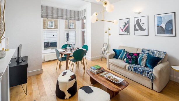 A modern living room with a sofa, coffee table, and ottoman. There is a dining area with chairs and a table. The room has artwork on the walls and a chandelier.