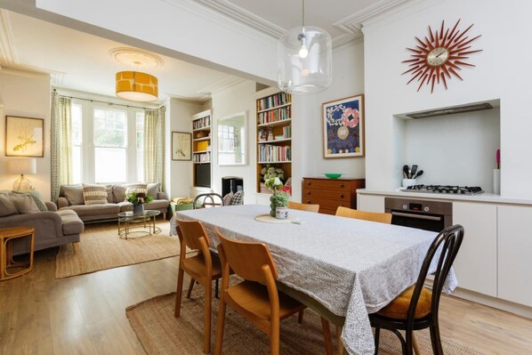 A modern kitchen with a dining area, featuring a large table, chairs, a hanging light fixture, and a clock on the wall. - Stunning 2 Bed Apt W Garden in Clapham (London)