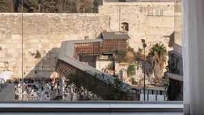 A view from a room with a large arched window overlooking a historic stone wall, a wooden structure, and a distant building with a tower. - Western Wall View Apartment (Jerusalem)