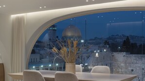 A modern dining area with a large table and chairs, a vase with dried plants, and a view of a historic site through a large window. - Western Wall View Apartment (Jerusalem)