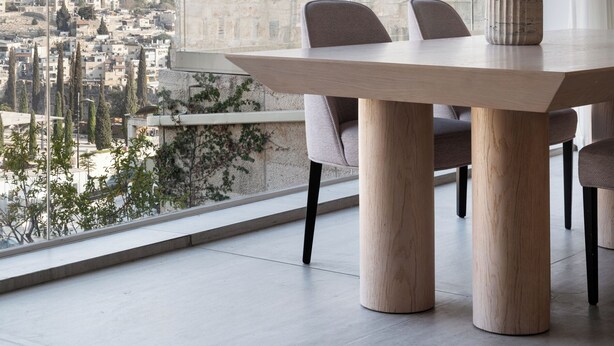 A modern dining area with a large table and chairs, a vase with dried plants, and a view of a cityscape through a large window. - Western Wall View Apartment (Jerusalem)