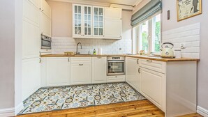 A kitchen with white cabinets, a tiled backsplash, a wooden floor, and a window with a view of greenery. - Victus Apartament Dębowa (Sopot)