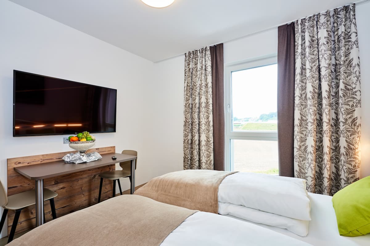 A hotel room with a bed, a desk with a bowl of fruit, a flat-screen TV, and a window with curtains.