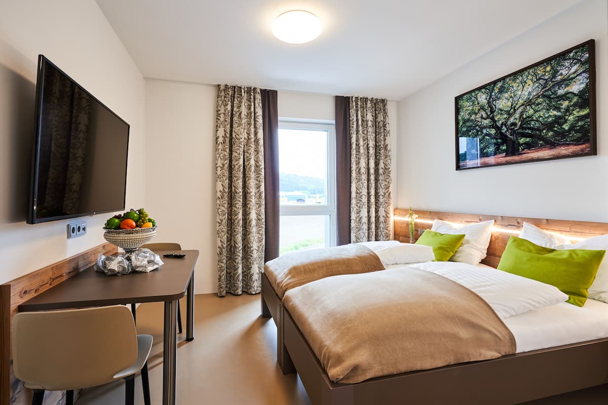 A hotel room with a bed, a desk with a fruit bowl, a TV, and a painting on the wall.
