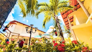 A courtyard with palm trees, a building with a red-tiled roof, and outdoor seating with umbrellas. - Pousada Paraíso das Palmeiras (Paripueira)