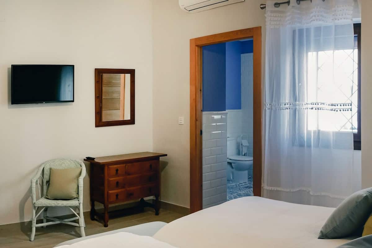 Une chambre à coucher avec un lit, une chaise, une commode, un téléviseur et une fenêtre avec un rideau.