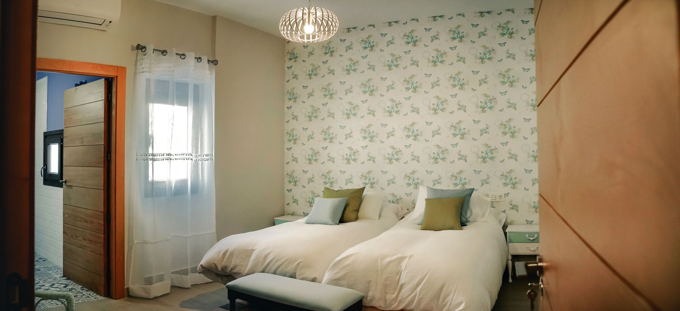 A bedroom with two beds, a wooden door, a window with sheer curtains, a patterned wallpaper, and a hanging light fixture.