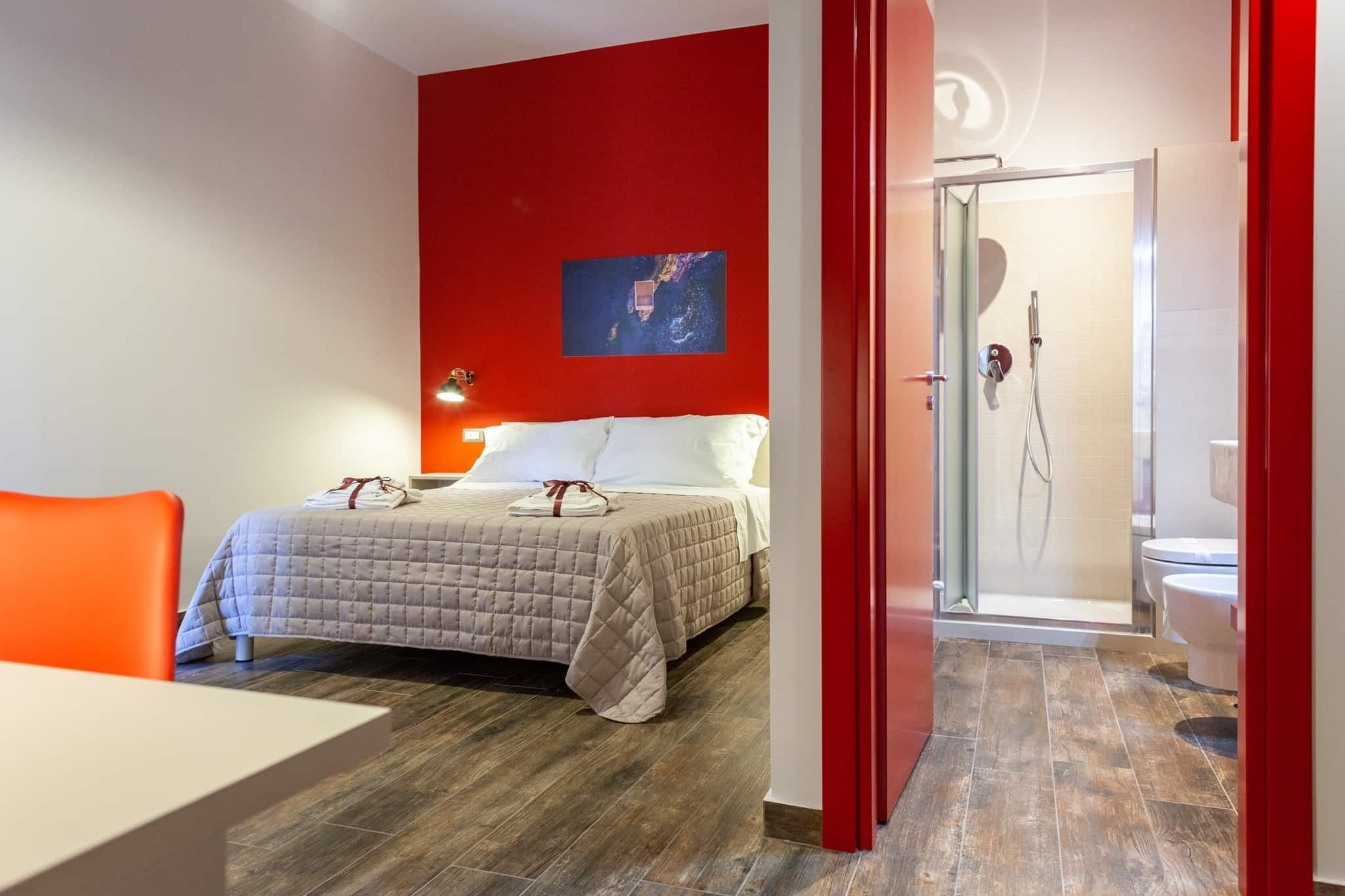 A hotel room with a bed, a red accent wall, a shower area, and a white toilet.