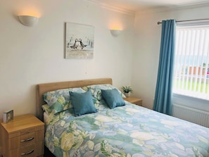 A bedroom with a floral bedspread, a wooden nightstand with drawers, a bedside table with a plant, and a window with curtains. - 3 bedroom accommodation in Aberdaron (Aberdaron)