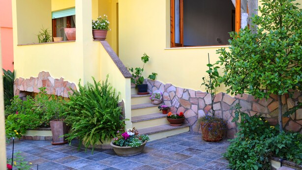 A house with a stone staircase leading to a balcony, surrounded by various potted plants and a small garden area.