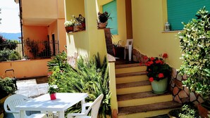 A sunny outdoor patio with a white table and chairs, potted plants, and a staircase leading to a green door.