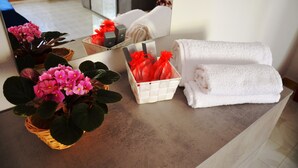 A bathroom counter with a mirror, a basket of pink flowers, a basket of red gift bags, and folded white towels.