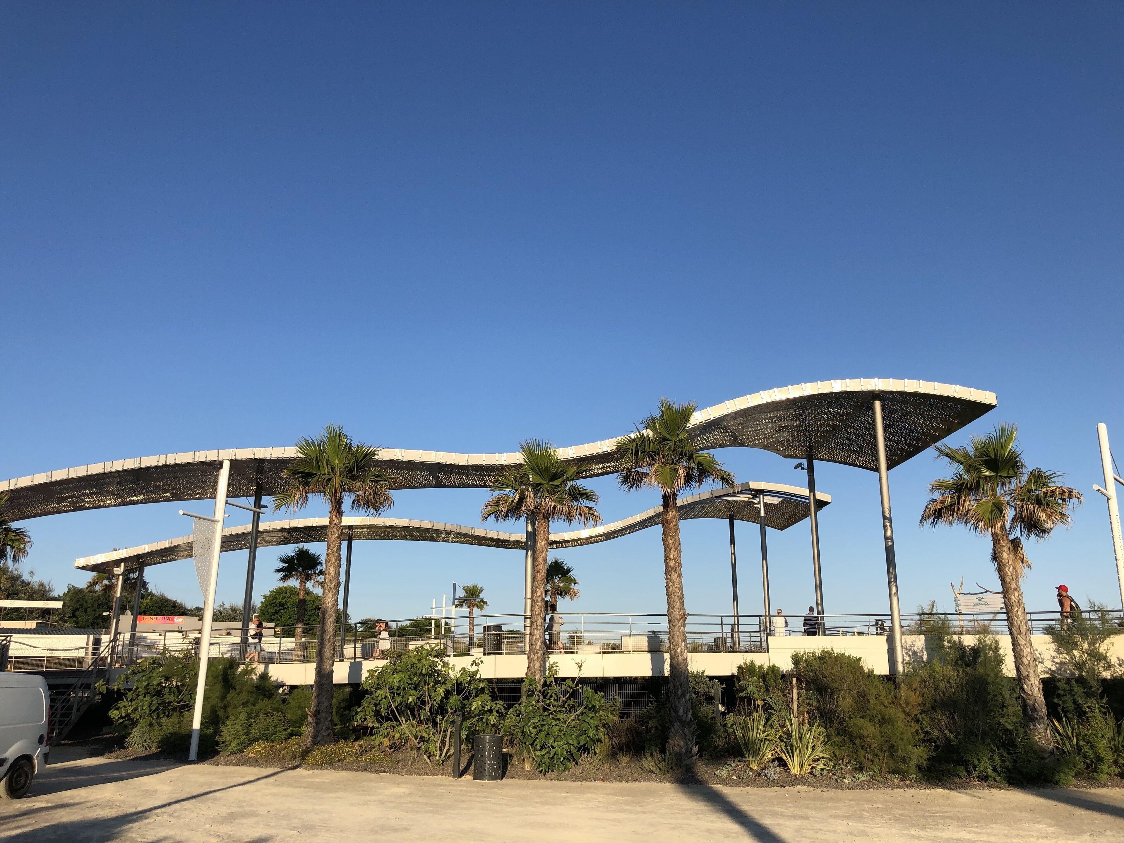 Un espacio exterior moderno con palmeras, una camioneta blanca y un puente peatonal.