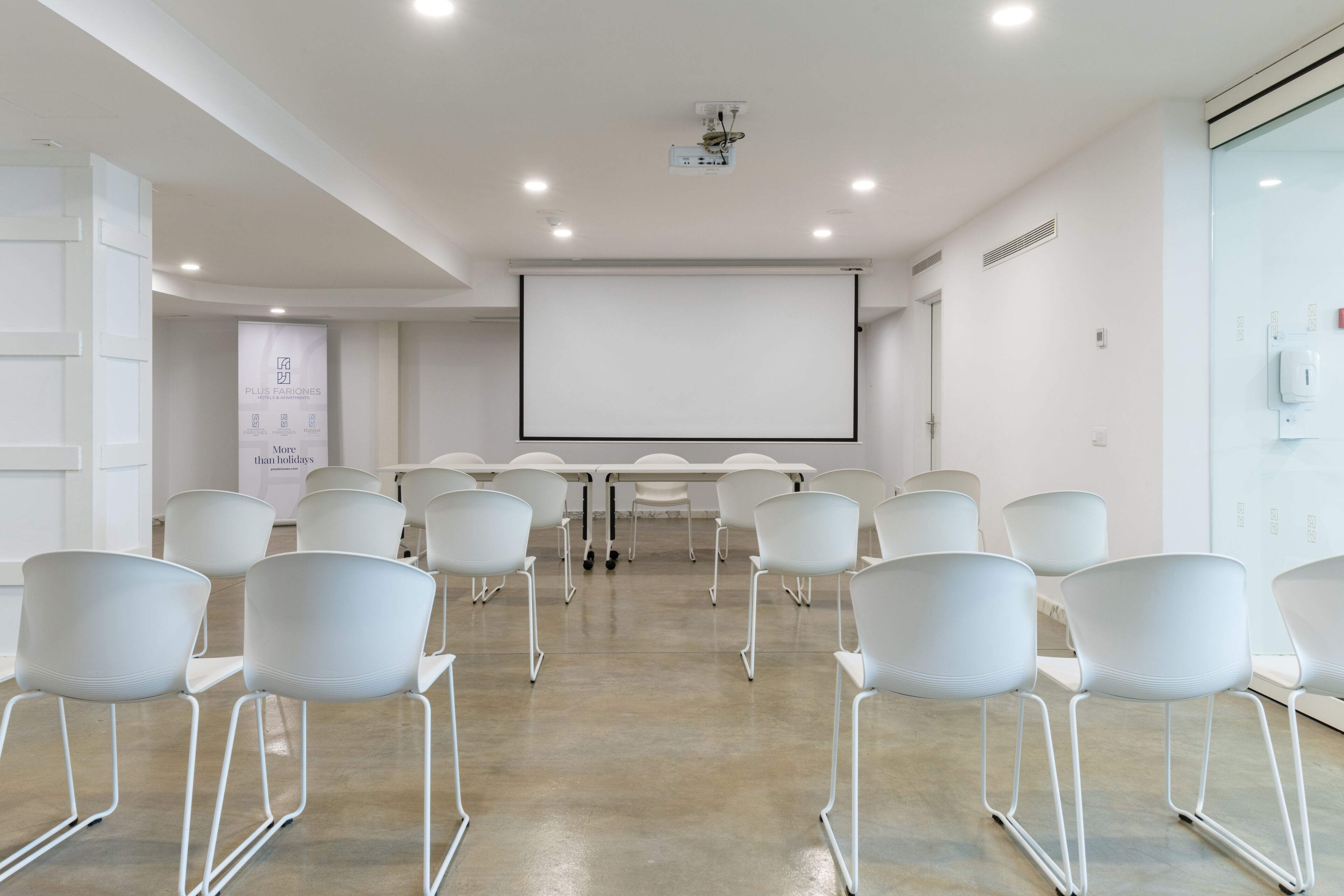 Una sala de conferencias con sillas blancas, una mesa grande, una pantalla de proyector y una pizarra.