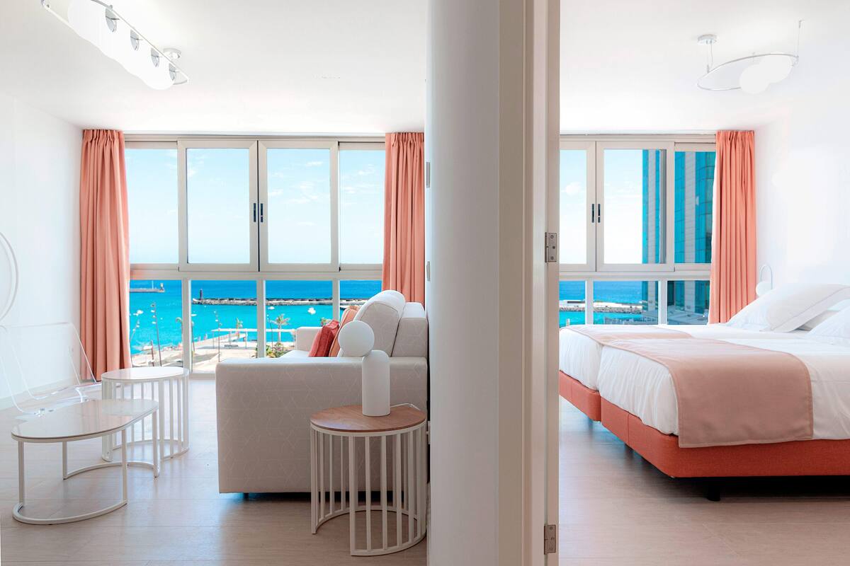 Ein modernes Hotelzimmer mit großem Fenster und Meerblick, einem Bett mit rosa Kopfteil, einem weißen Sofa und einem kleinen runden Tisch.