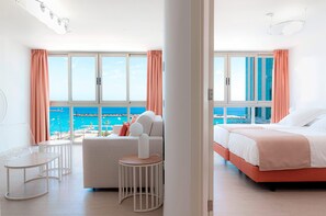 A modern hotel room with a large window offering a view of the sea, a bed with a pink headboard, a white sofa, and a small round table. - Plus Fariones Habitat (Arrecife)