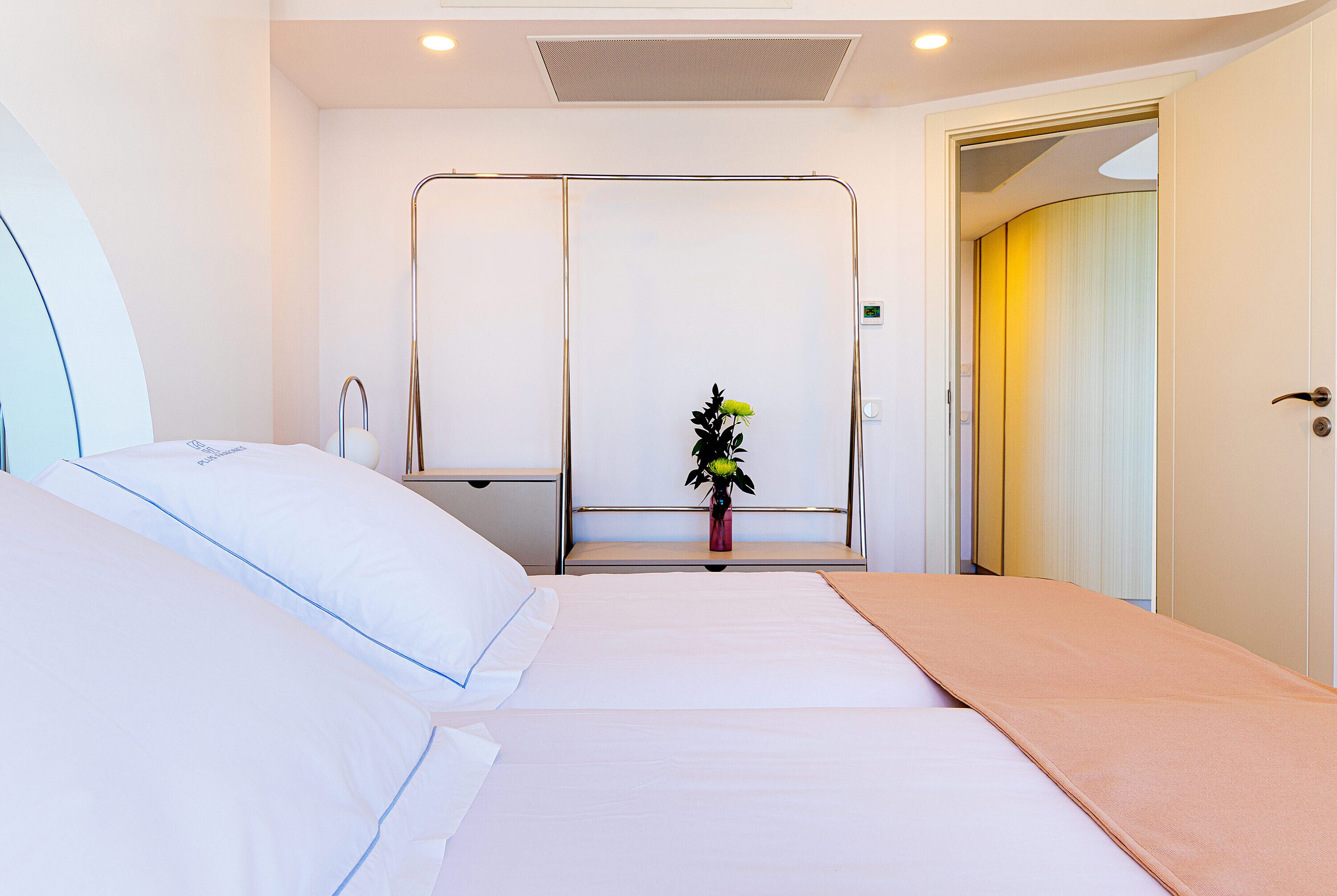 Une chambre d’hôtel comprenant un lit, une table de chevet, une petite table d’appoint, une petite plante en pot et une porte donnant sur une autre pièce.