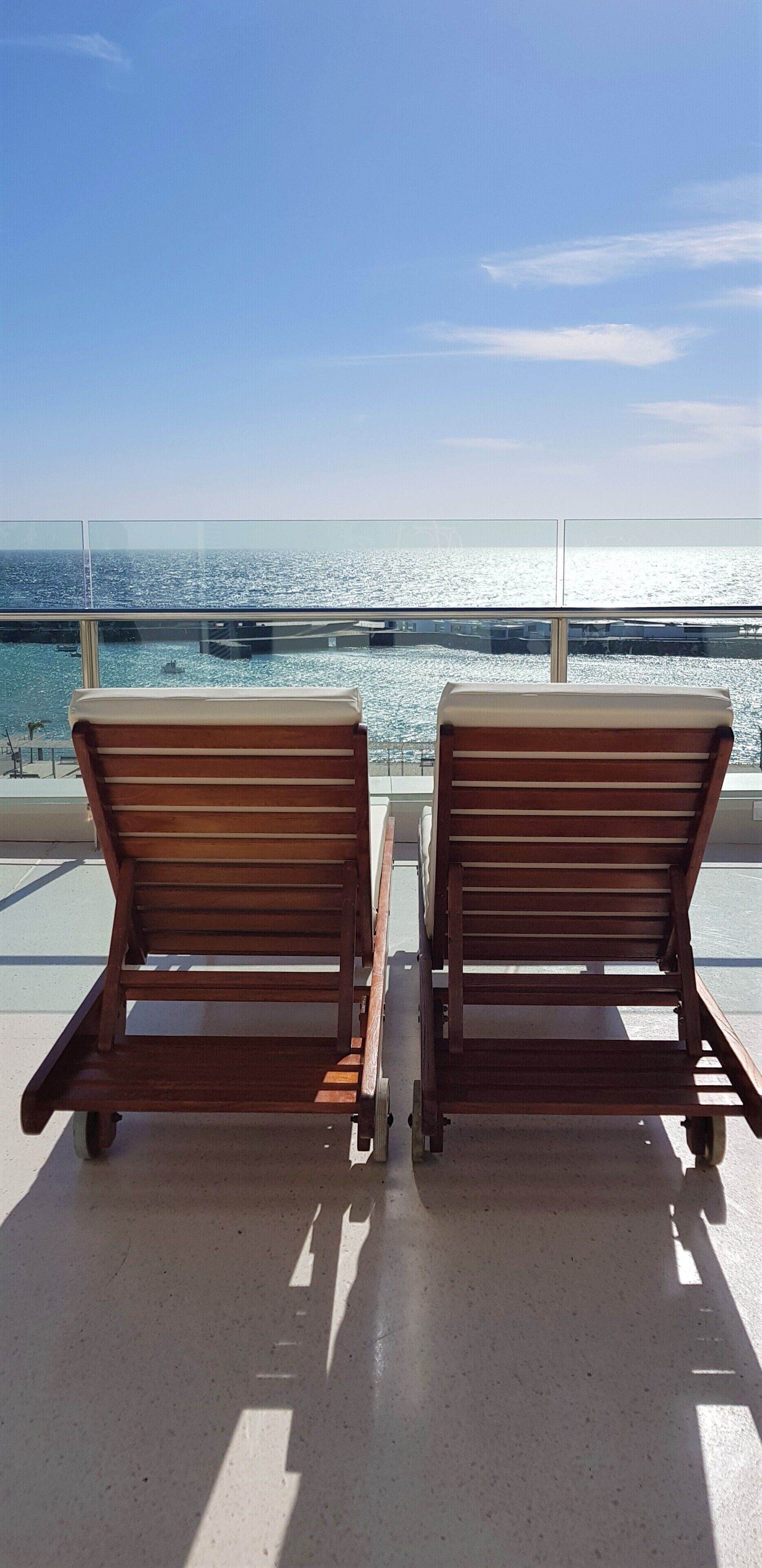 Two wooden lounge chairs with wheels on a balcony overlooking a body of water.