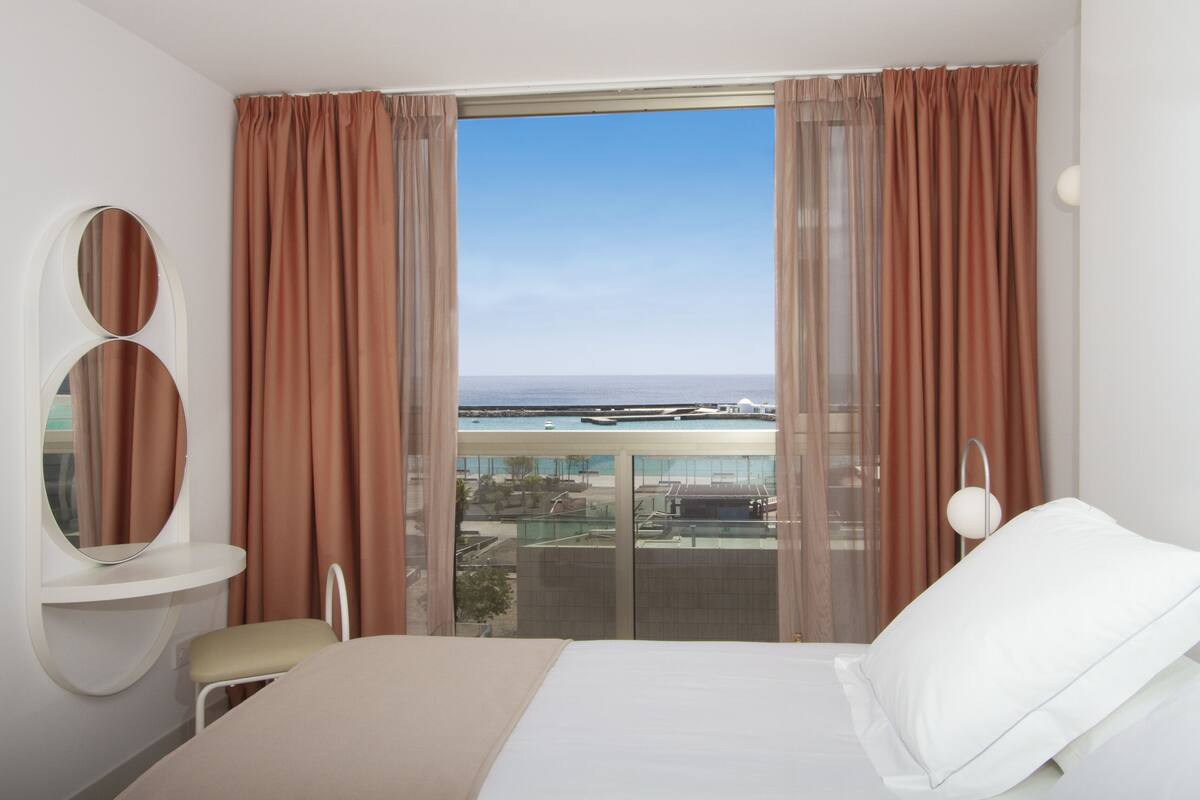 Ein Hotelzimmer mit einem Bett, einem Sessel, einem runden Spiegel und Blick auf einen Yachthafen durch große Fenster.