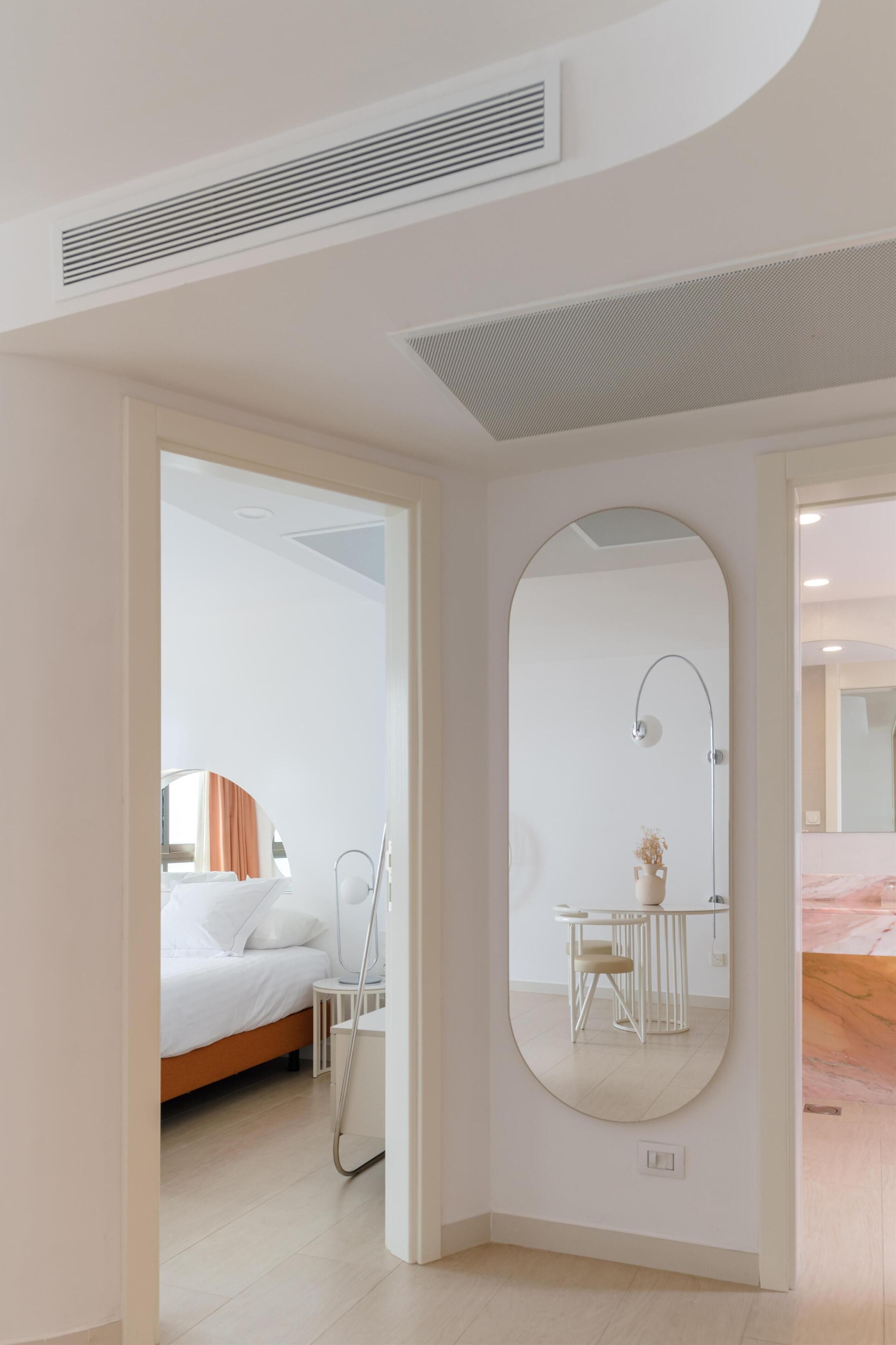 A modern hotel room with a large mirror, a bed, a bedside table, and a small seating area.