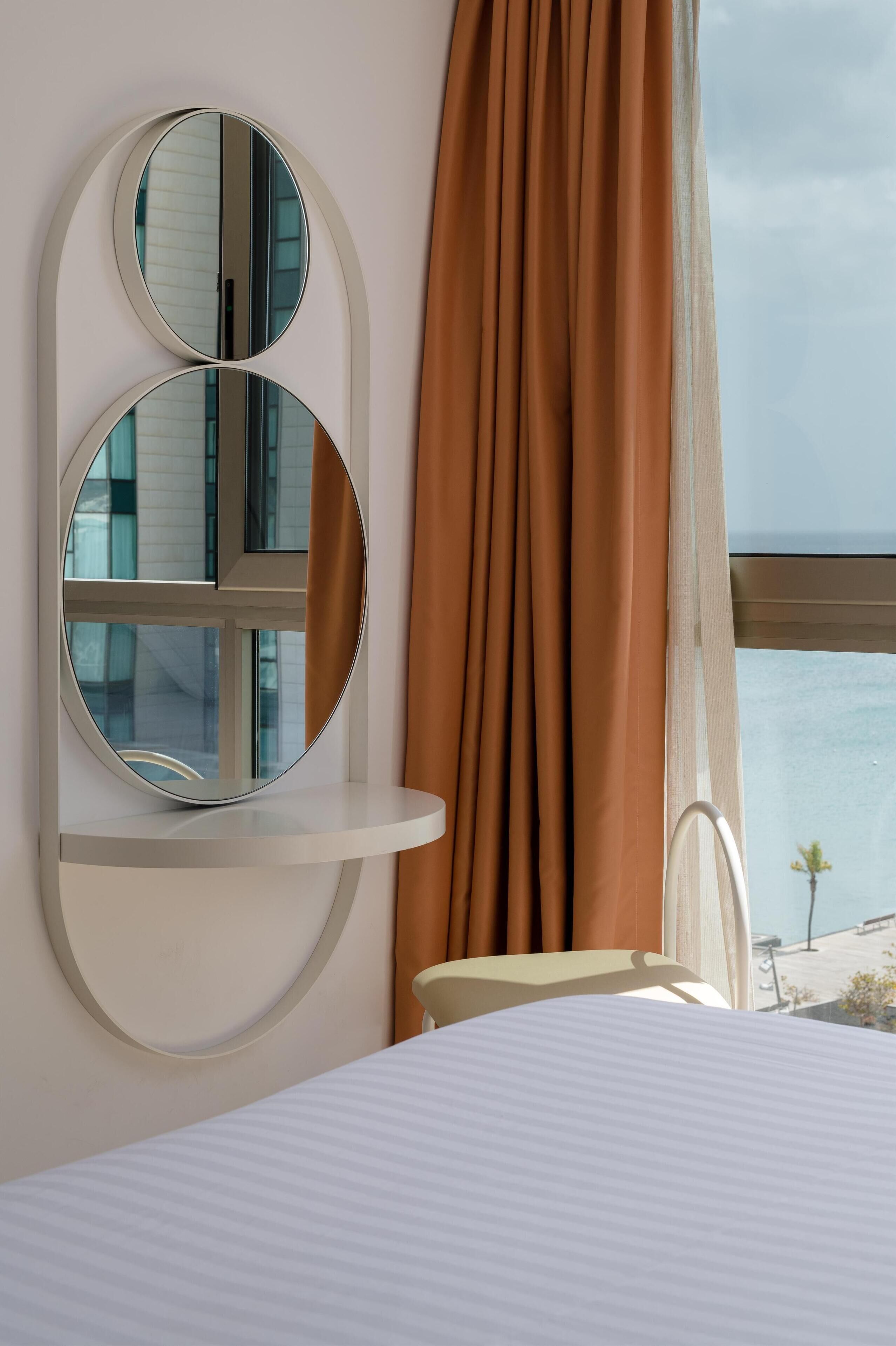 A bedroom with a bed, a round mirror, and a view of the sea through a window with curtains.