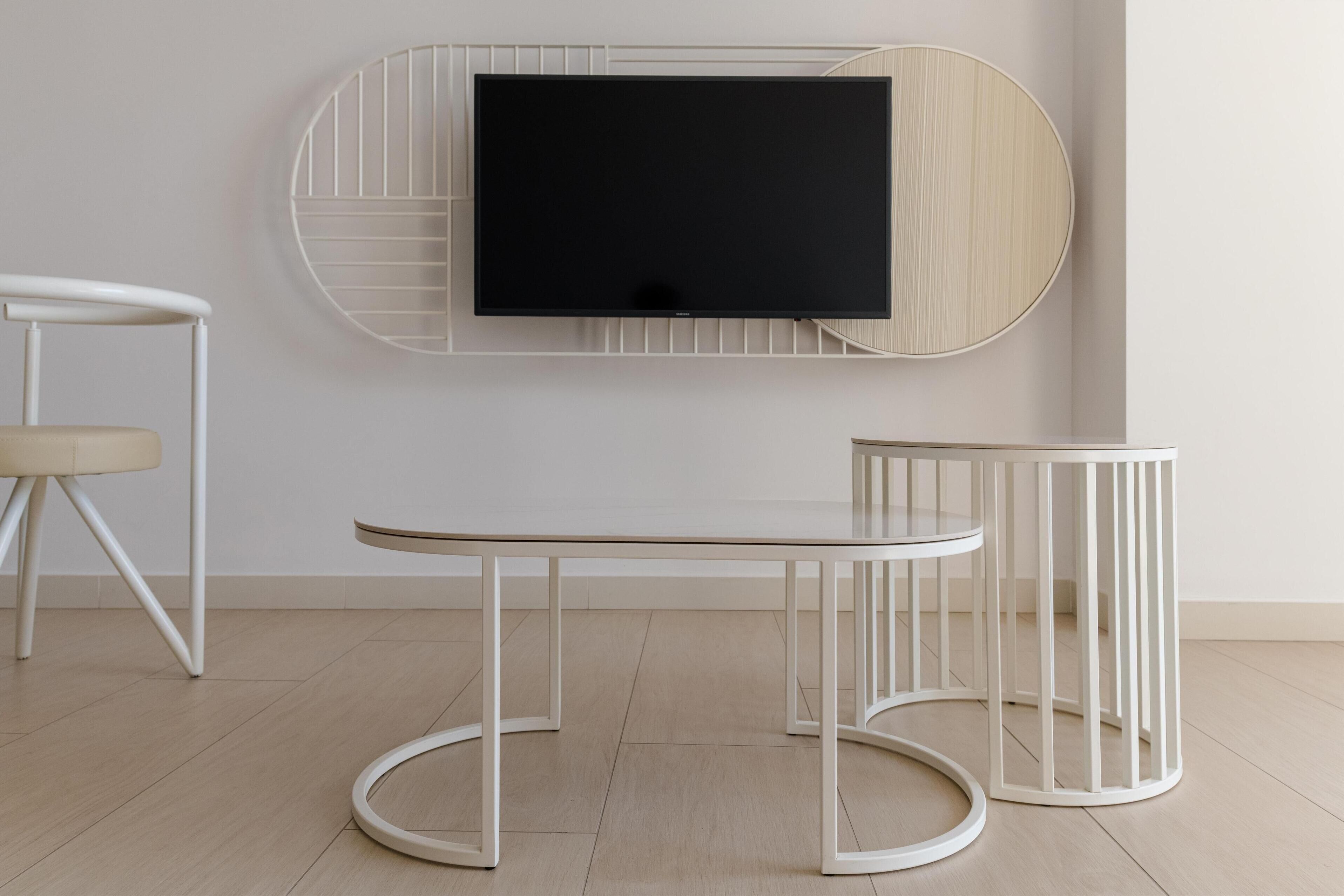A minimalist room with a flat-screen TV mounted on a wooden panel, a white table with a circular base, and a white chair with a cushioned seat.
