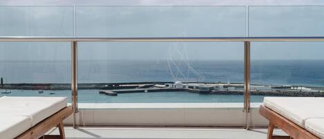 A balcony with two wooden beds and a glass railing overlooking a marina with boats and a pier.