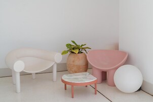 Una habitación minimalista con una silla blanca, una mesita pequeña, un taburete rosa, una planta en maceta y un objeto esférico blanco.