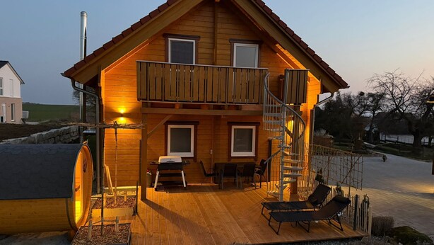 A two-story wooden house with a balcony, outdoor seating, and a wooden deck.