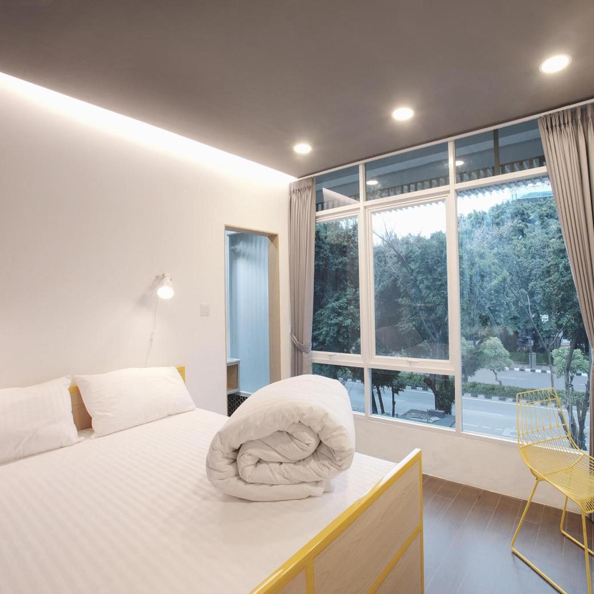 A hotel room with a bed, a window with curtains, a chair, and a wall-mounted light.