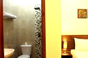 A bathroom with a sink, toilet, and shower. There is a towel rack with a towel hanging on it. The bathroom has a checkered tile backsplash and a wooden headboard with a lamp beside the bed. - Grand Marto Hotel (Yogyakarta)