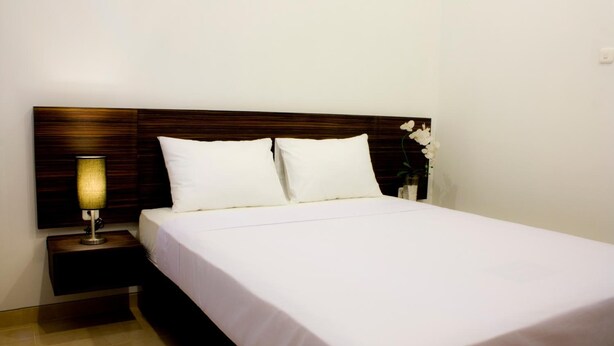 A neatly made bed with white linens, a wooden headboard, a bedside lamp, and a small vase with white flowers. - Grand Marto Hotel (Yogyakarta)