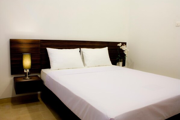 A neatly made bed with white linens, a wooden headboard, a bedside lamp, and a small vase with white flowers. - Grand Marto Hotel (Yogyakarta)