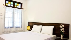 A neatly made bed with white linens, a wooden headboard, a nightstand with a lamp, and a window with a stained glass design. - Grand Marto Hotel (Yogyakarta)