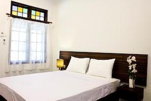 A neatly made bed with white linens, a wooden headboard, a nightstand with a lamp, and a window with a stained glass design. - Grand Marto Hotel (Yogyakarta)