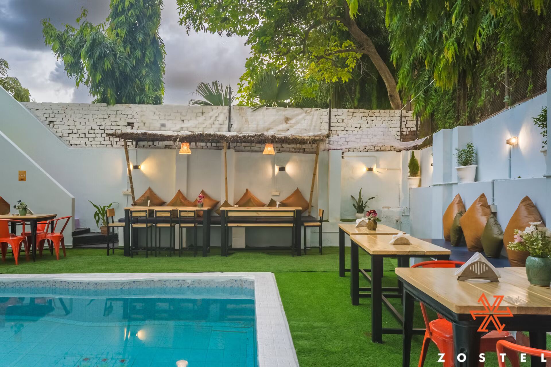 An outdoor dining area with tables and chairs, a pool, and a brick wall with plants.
