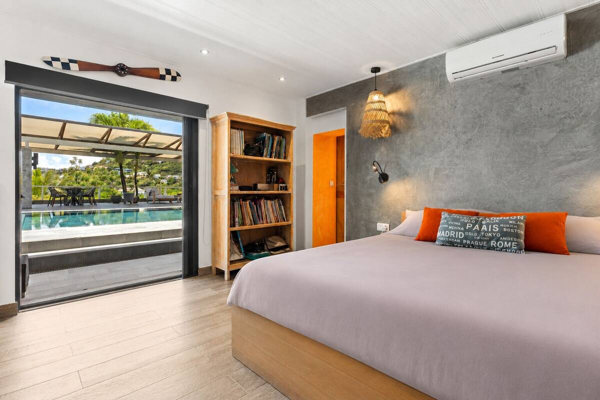 A bedroom with a large bed, a bookshelf, an air conditioning unit, and a view of an outdoor swimming pool.