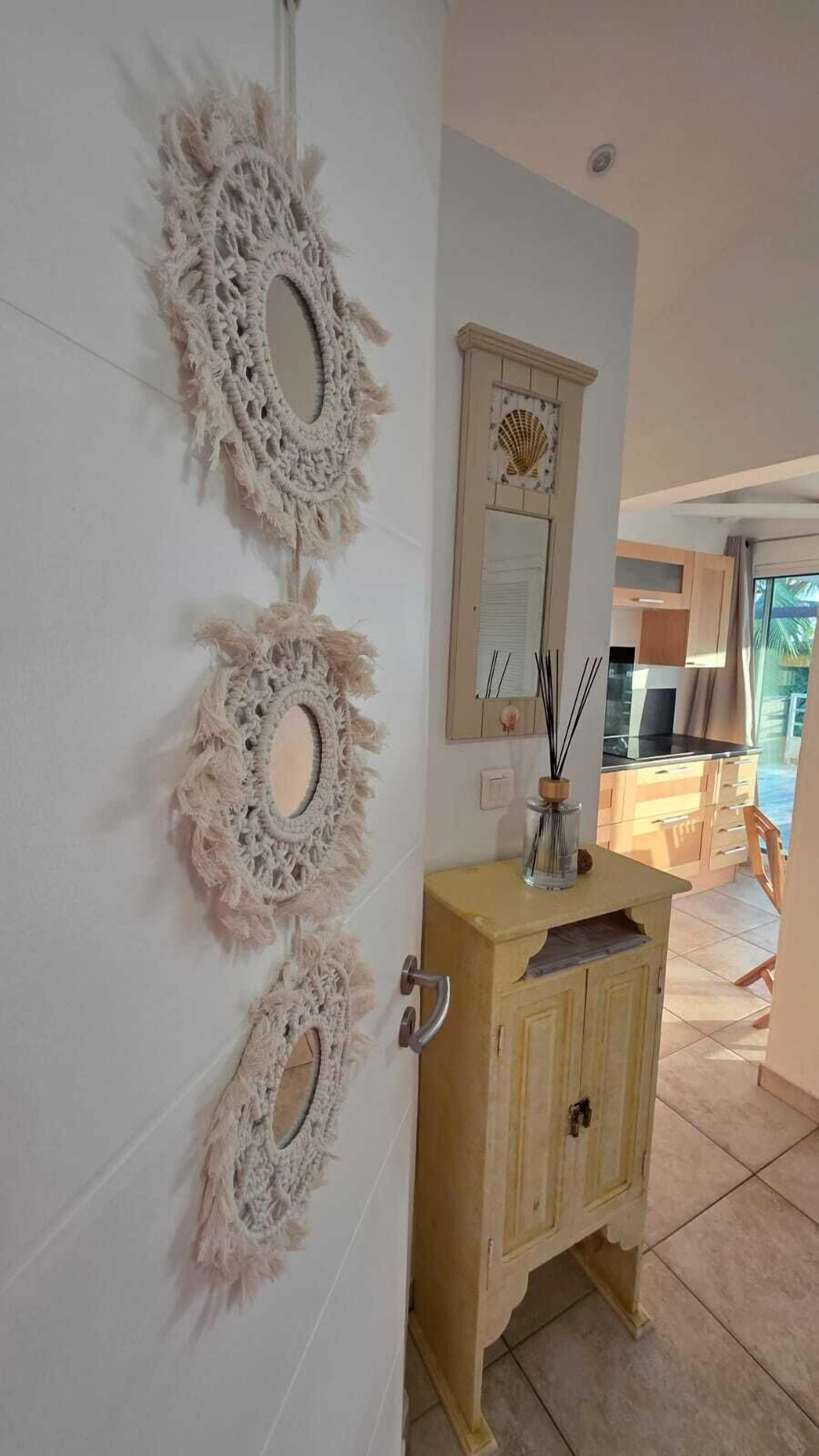 Um quarto com uma porta decorada com três peças de macramé, um armário de madeira com um espelho em cima e vista para uma cozinha e área externa através de uma porta de vidro.