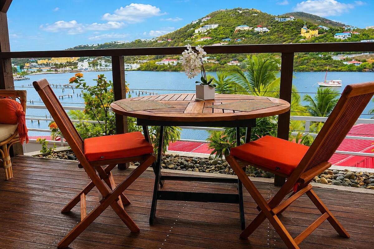 A wooden deck with a round table and two chairs, overlooking a body of water and a hillside with buildings.
