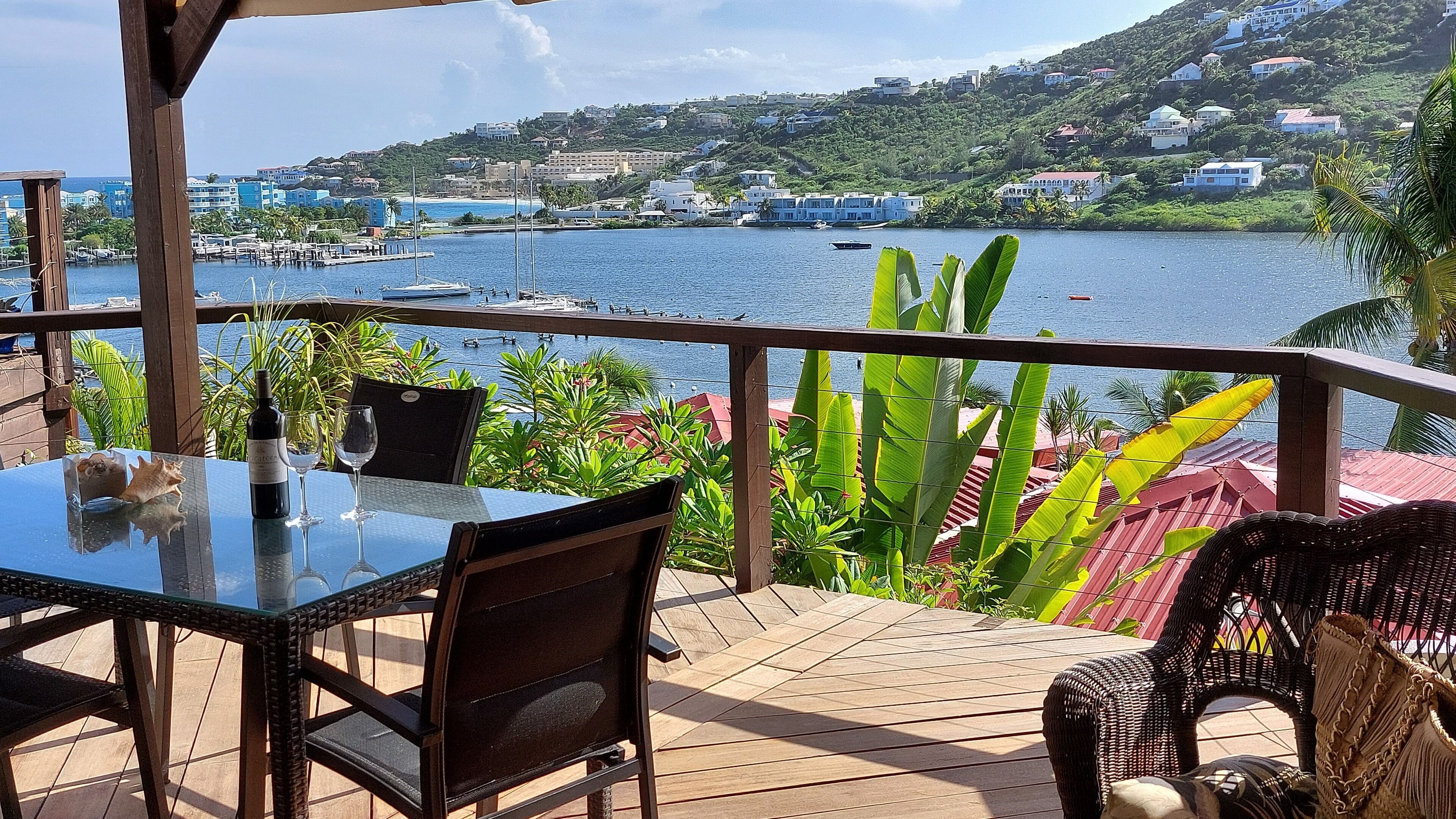 Une terrasse avec une table et des chaises, donnant sur une marina où sont amarrés des bateaux, et sur un paysage vallonné en arrière-plan.