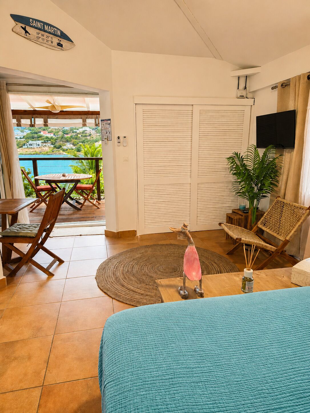 A room with a view of a coastal landscape, a wooden table with chairs, a large bed, a TV mounted on the wall, and a sliding door.