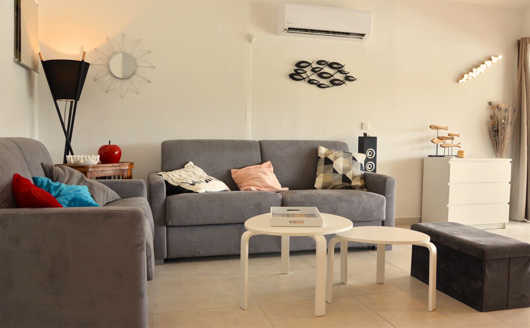 A living room with a grey sofa, a small white table, a black ottoman, a red apple, and a decorative wall piece.