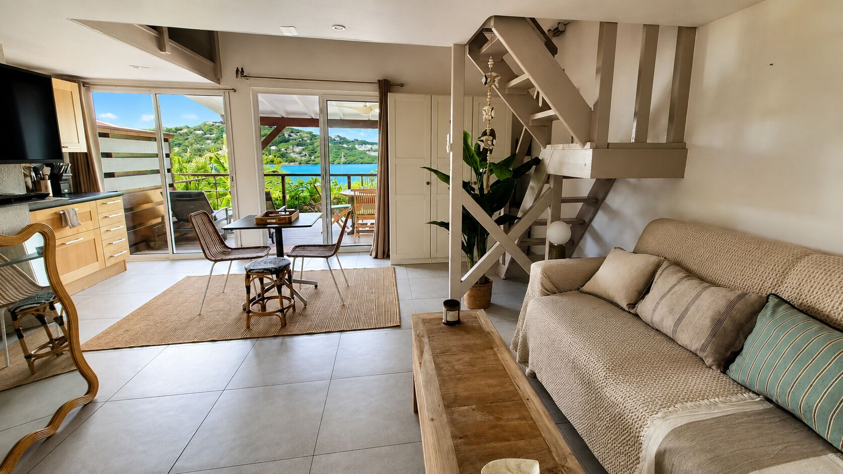 A spacious living area with a sofa, dining table, and chairs, a kitchen area, and a staircase leading to a balcony with a scenic view.