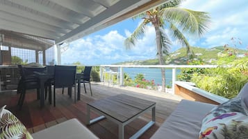 A covered outdoor seating area with a wooden table and chairs, a cushioned sofa, and a view of the sea and hills.