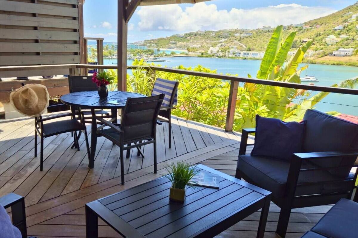 A wooden deck with outdoor furniture, including a table and chairs, overlooking a scenic view of a body of water and hills.