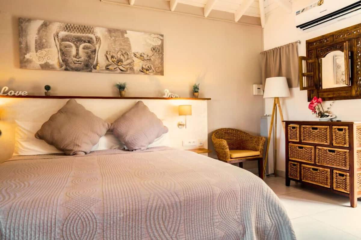 A bedroom with a large bed, a headboard with a shelf, a wall-mounted Buddha painting, a bedside lamp, a wicker chair, and a wooden dresser with baskets.