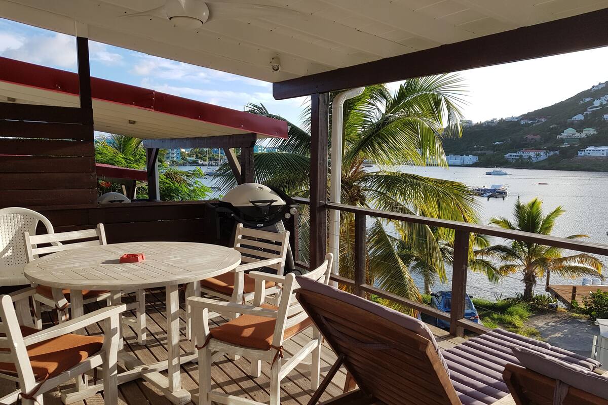 A balcony with a table and chairs, overlooking a body of water and a hillside.