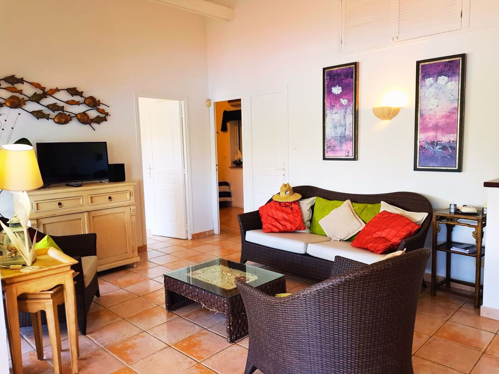 A living room with a sofa, coffee table, and a television on a cabinet. There are two framed artworks on the wall and a lamp on a side table.