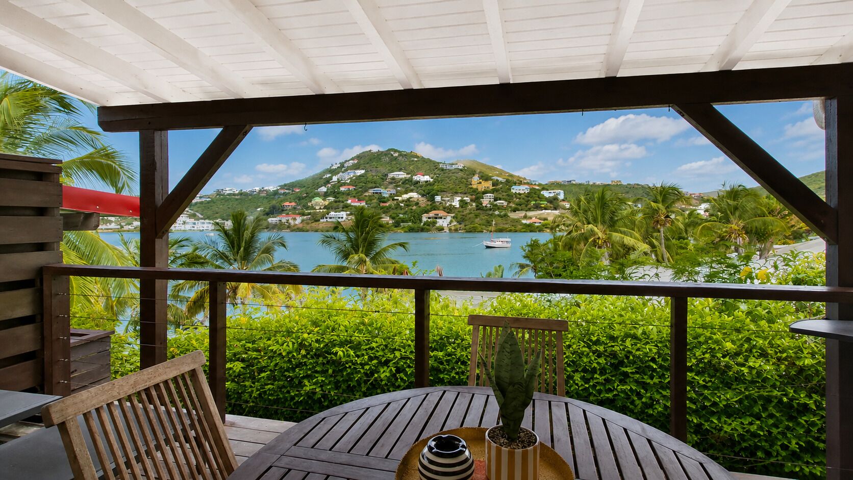 A covered outdoor area with a table and chairs, overlooking a body of water and a hillside with buildings.