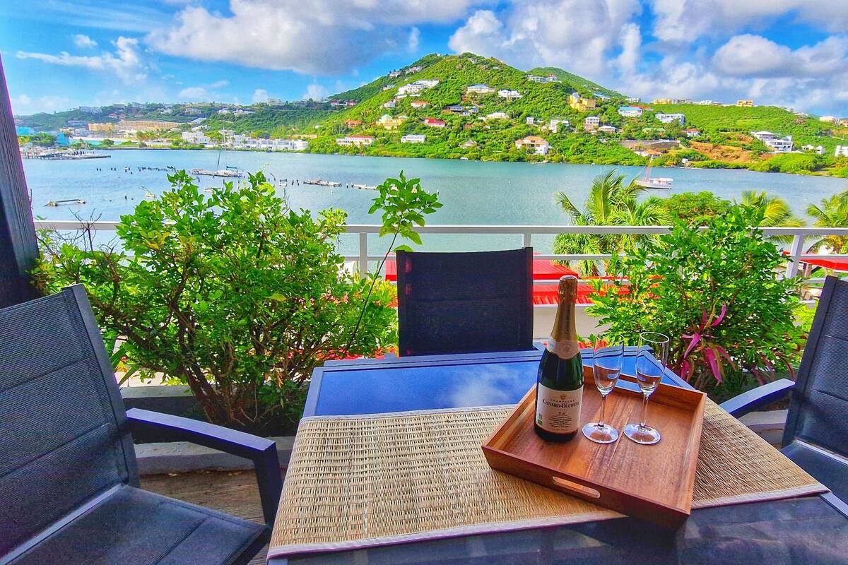 A balcony with two chairs, a table, and a bottle of champagne with two glasses on a tray. The balcony overlooks a scenic view of a lake with boats and houses on a hillside.